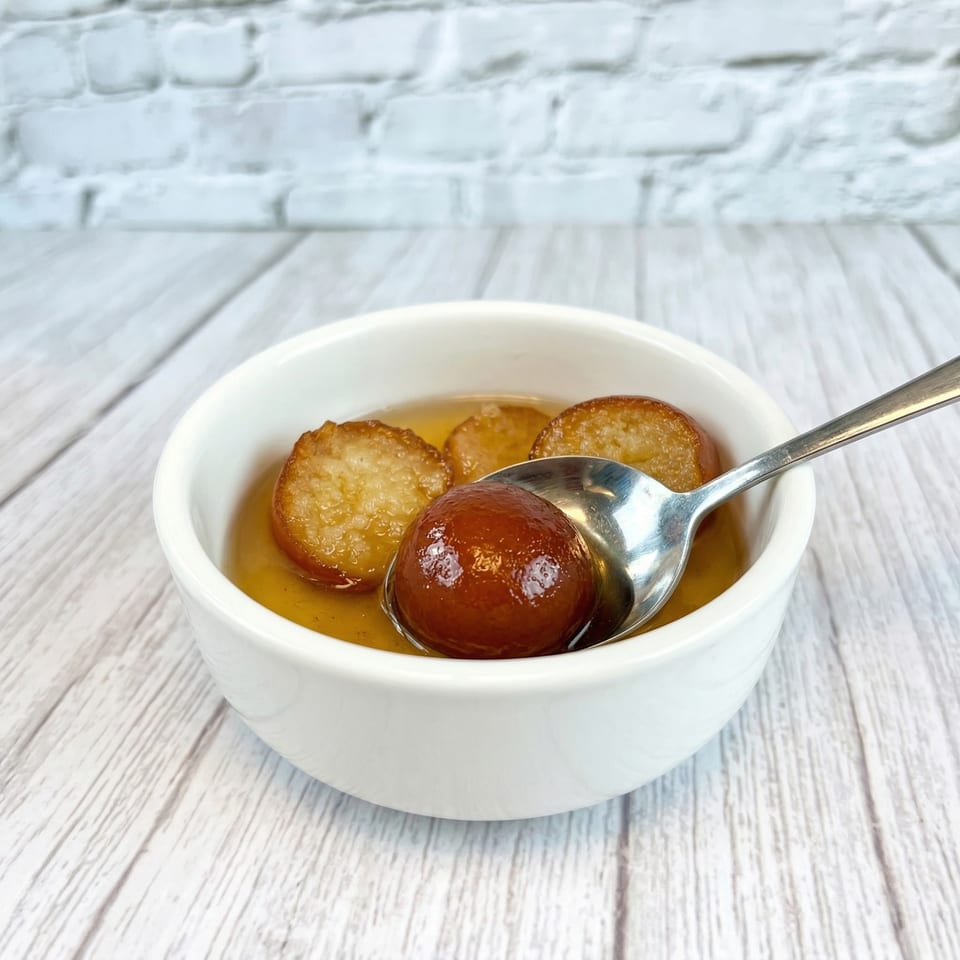 A white bowl of Gulab Jamun, with a spoon scooping one, on a light wooden table.