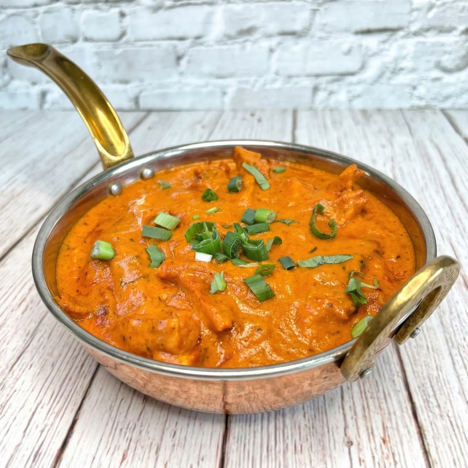A copper karahi filled with vibrant orange Indian butter chicken, garnished with fresh green onions.