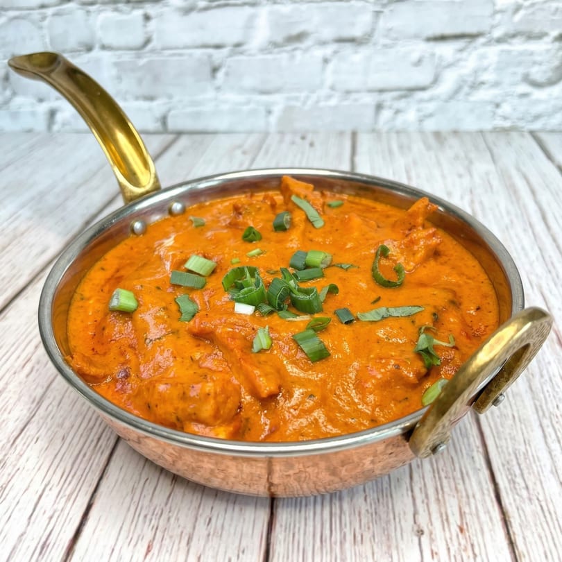 A copper karahi filled with vibrant orange Indian butter chicken, garnished with fresh green onions.