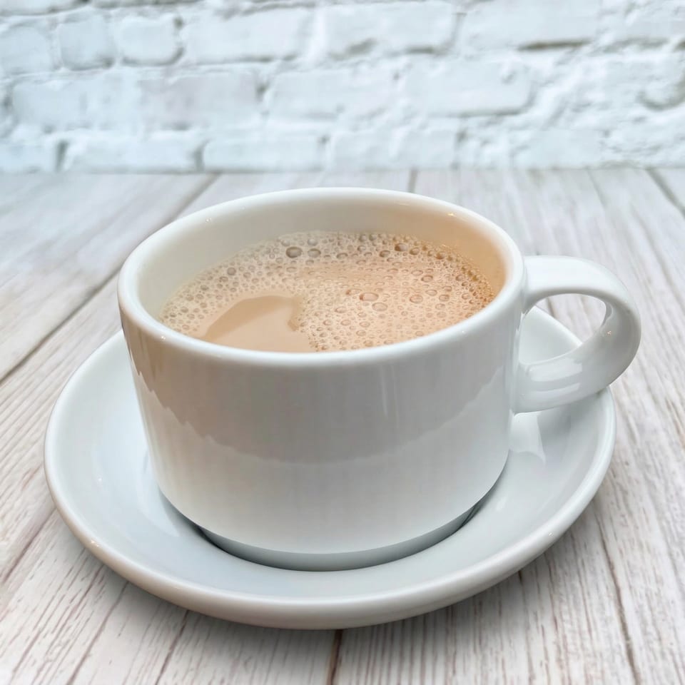 A white cup of Indian chai tea with foam, on a white saucer and wooden table.