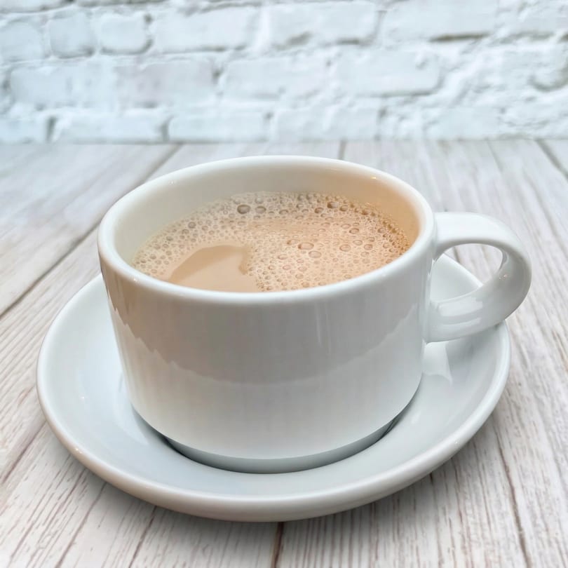 A white cup of Indian chai tea with foam, on a white saucer and wooden table.